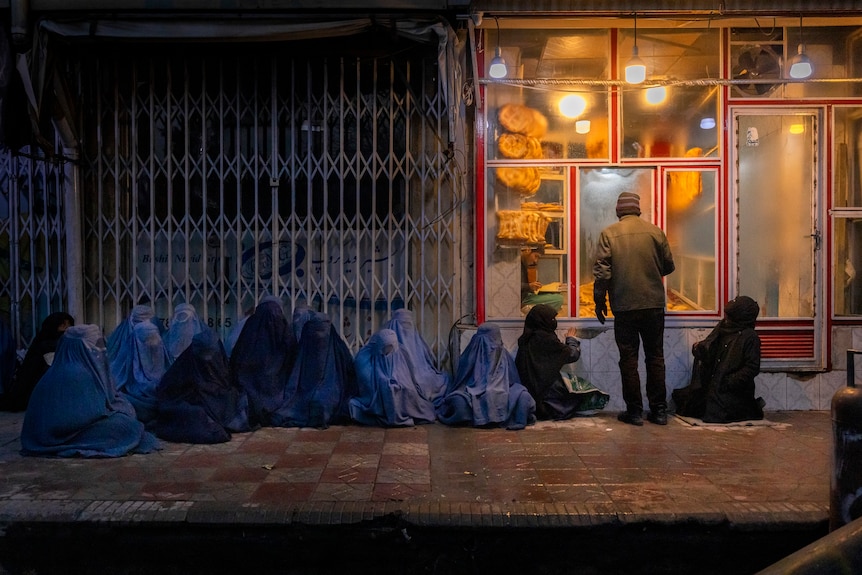 A man stands at the window of a lit-up bakery as 15 burqa-wearing women and children sit and kneel on the pavement outside.