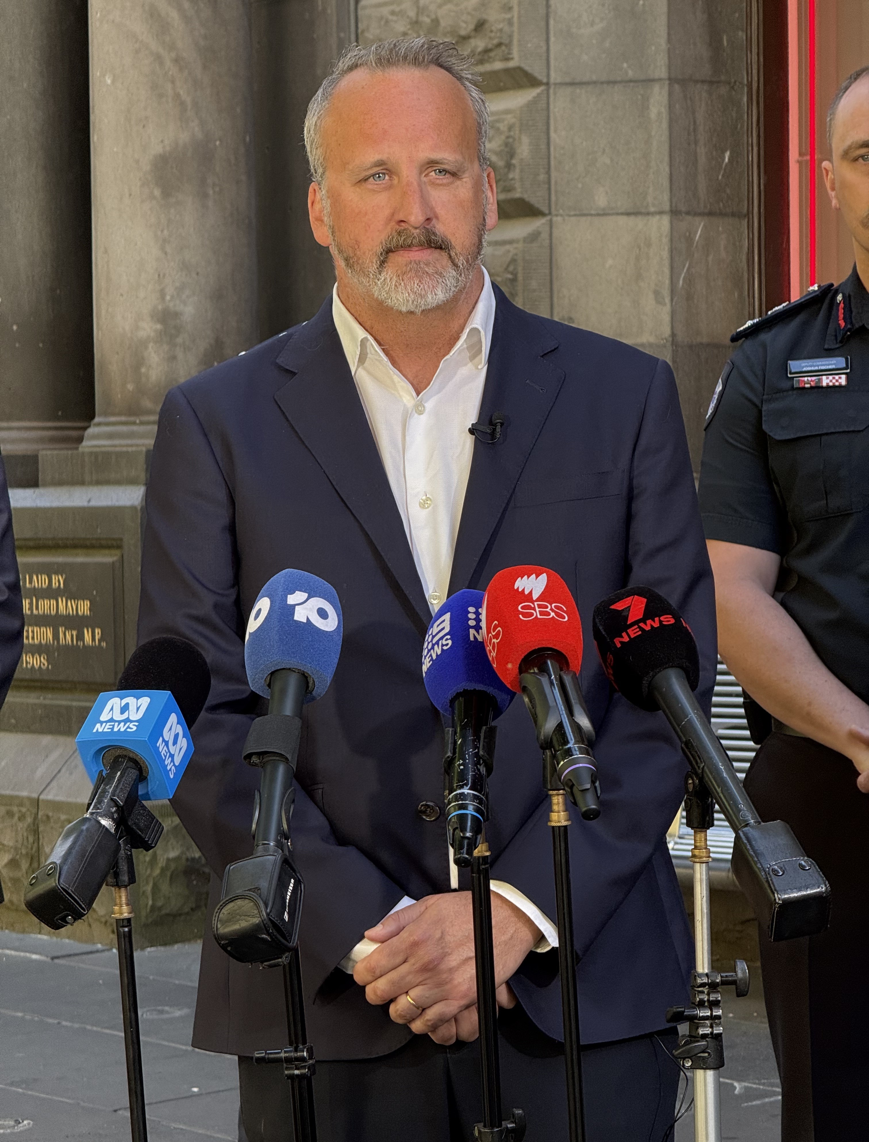 Um homem de cabelos grisalhos, bigode e barba, vestindo uma jaqueta azul marinho e camisa de colarinho branco, está atrás dos microfones.