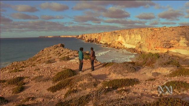 Two people stand on clifftop with ocean in the background