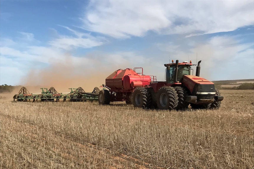 Red tractor pulling red air-seeder and green bar through stubble in a cloud of dust on sunny day