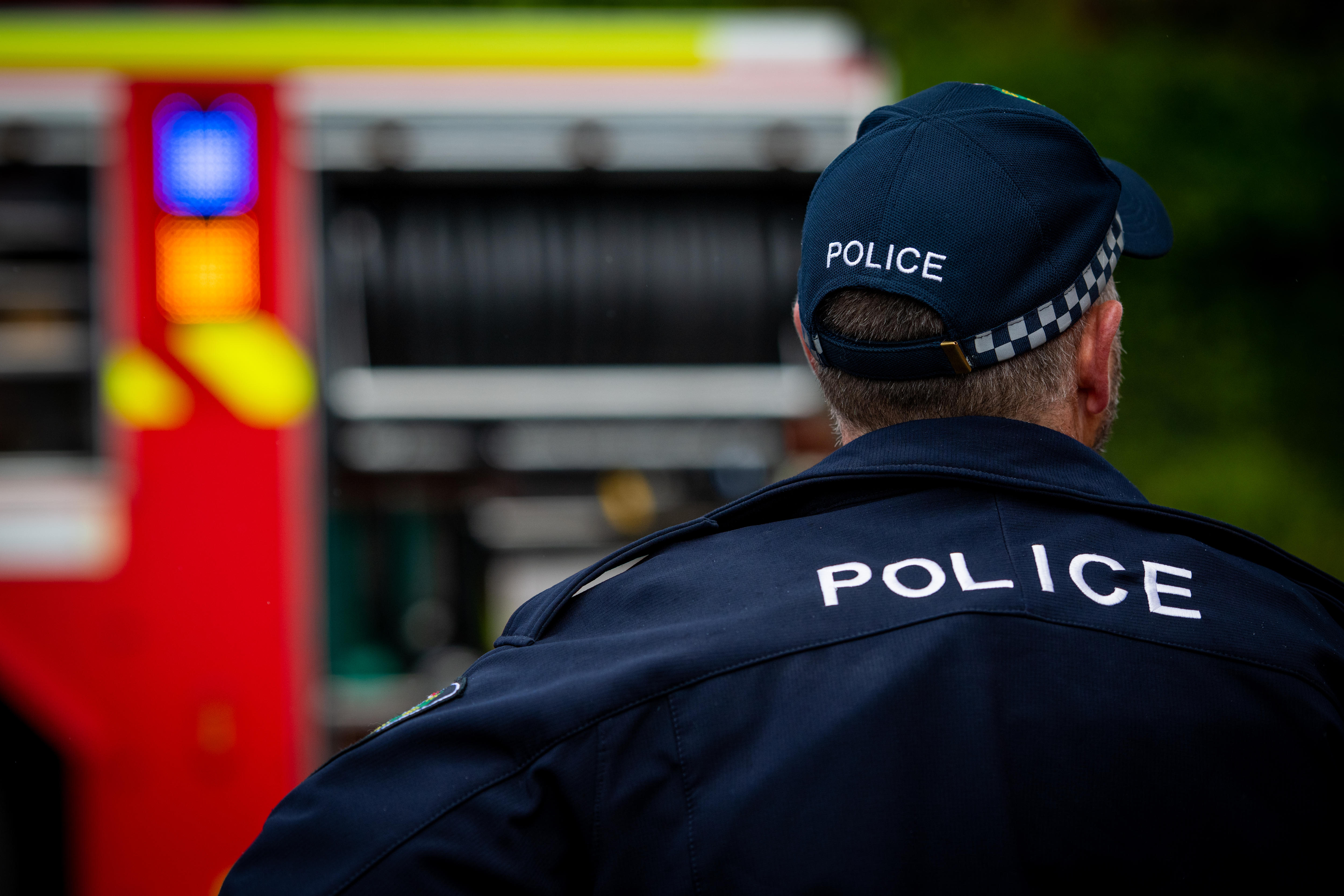 The back of a police officer wearing a blue police jacket and cap, with a red fire truck in the background