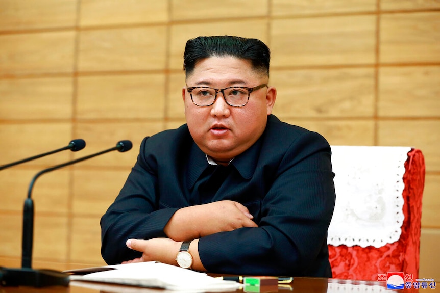 Kim Jong-un sits behind a mahogany desk and a bright red chair as he looks past two small microphones.
