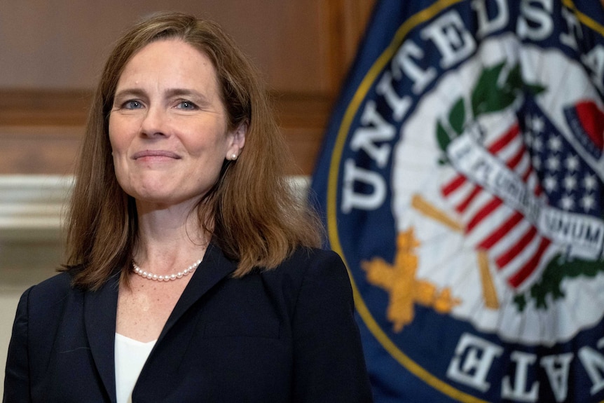 Amy Coney Barrett smiles in front of a flag saying 'United States'