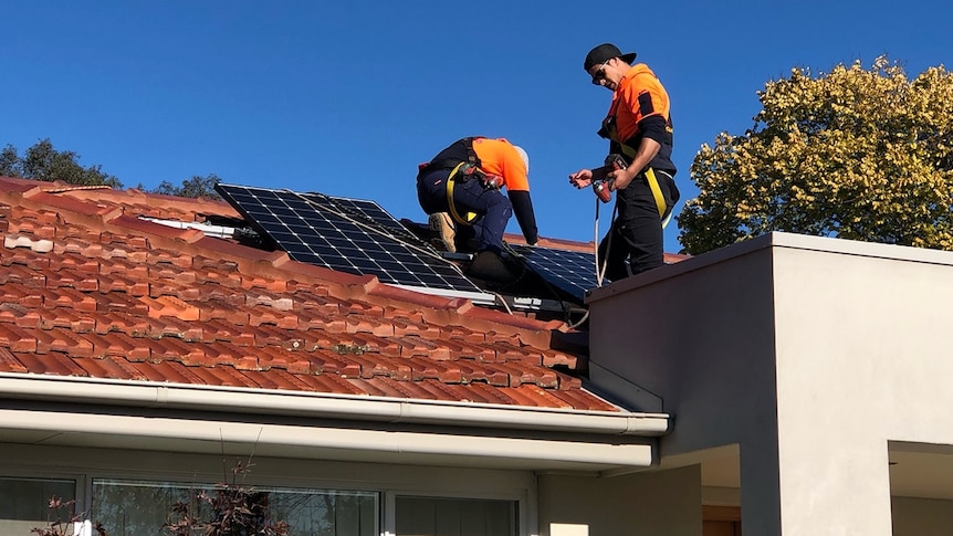 Two workers in safety gear install solar panels on the roof of a home.