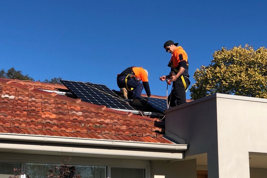 Two workers in safety gear install solar panels on the roof of a home.