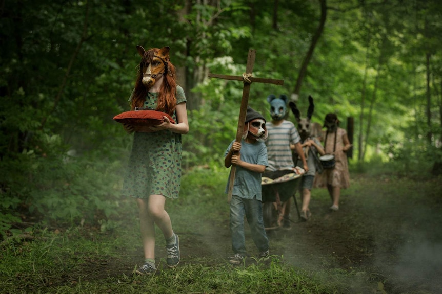 Walking in a line through a green wooded area, a procession of children wear unsettling animal masks and hold strange objects.