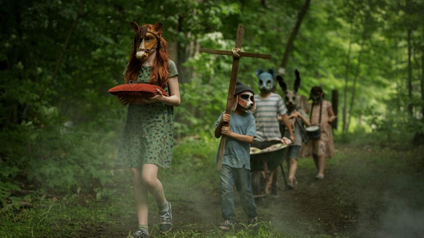 Walking in a line through a green wooded area, a procession of children wear unsettling animal masks and hold strange objects.