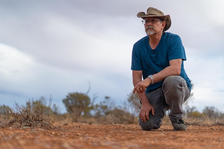 A man in a blue shirt and wide brimmed hat kneels on arid land.