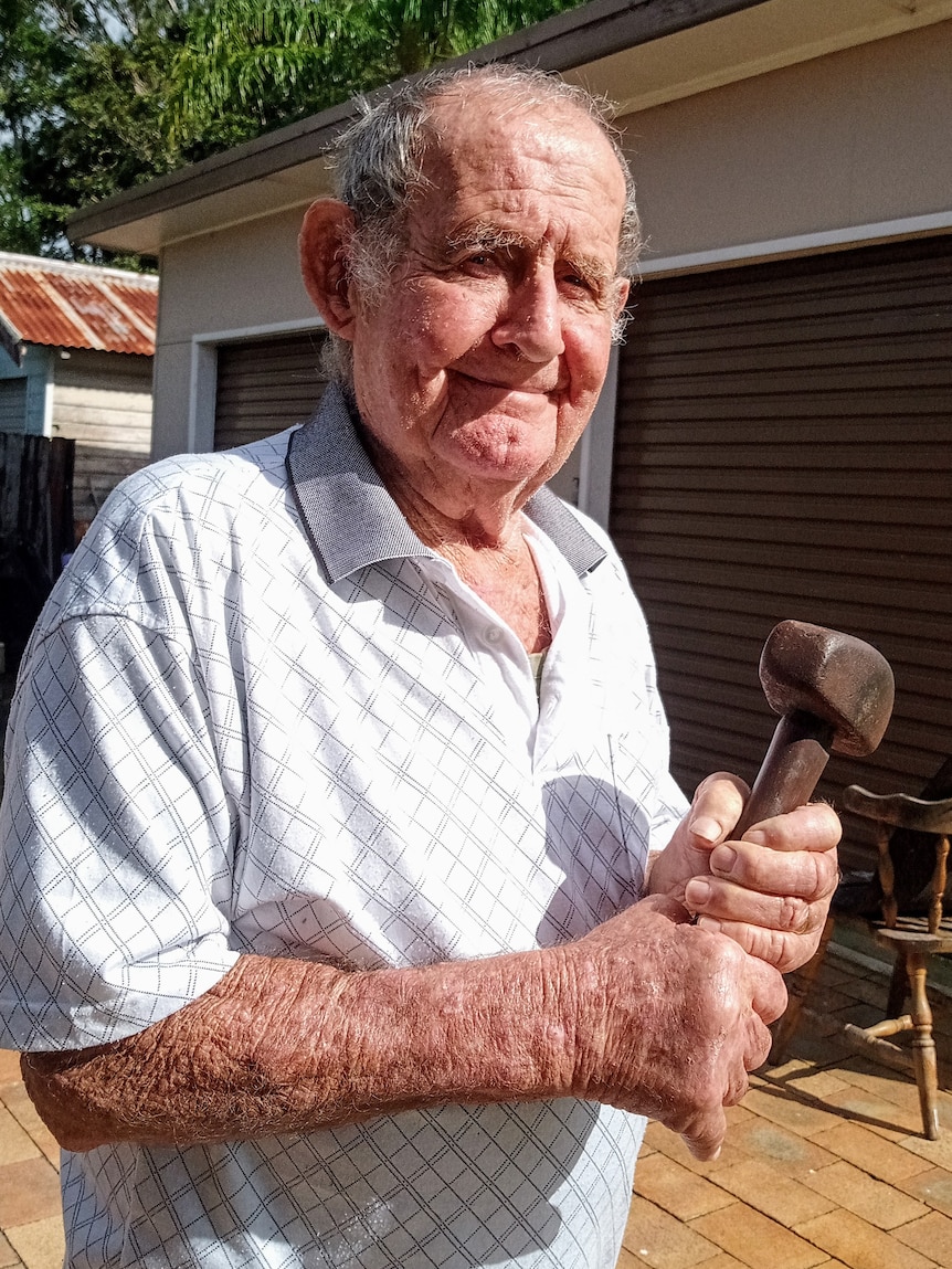 An elderly man in a light-coloured shirt holds a small, old hammer.