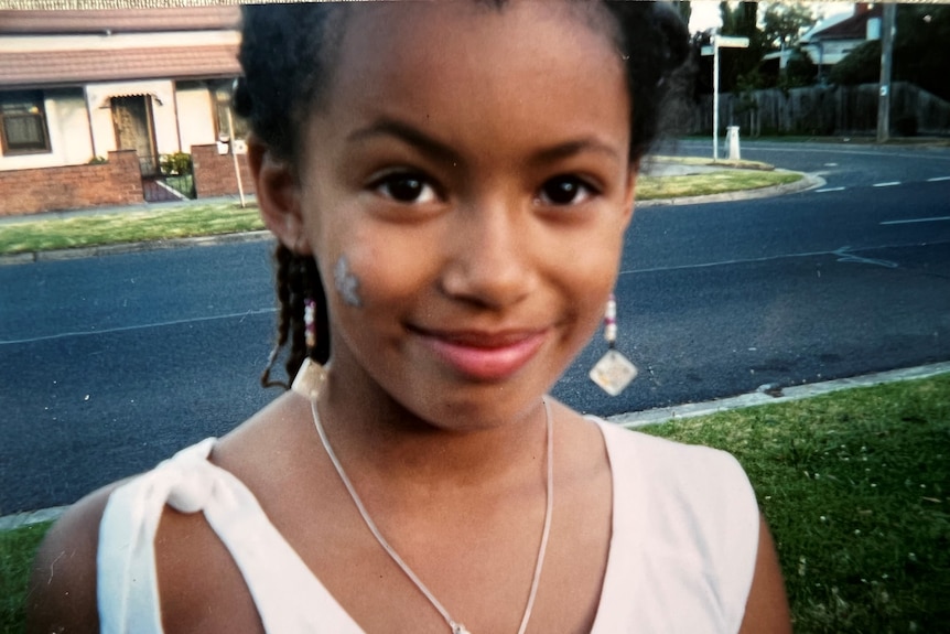 Yasmin Jeffery as a child looks to the camera with a faint smile. She wears a singlet, her hair is braided.