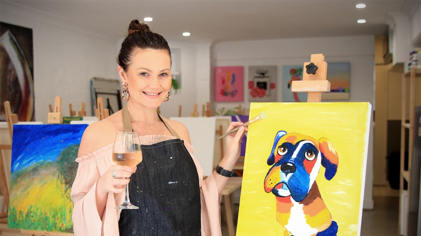 Brooke Hughes holds a wine and paintbrush standing next to an easel with an artwork of a dog.