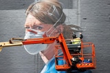 The subject of a mural is a woman wearing a PPE gown, face mask and goggles. A man continues painting the image on a grey wall.