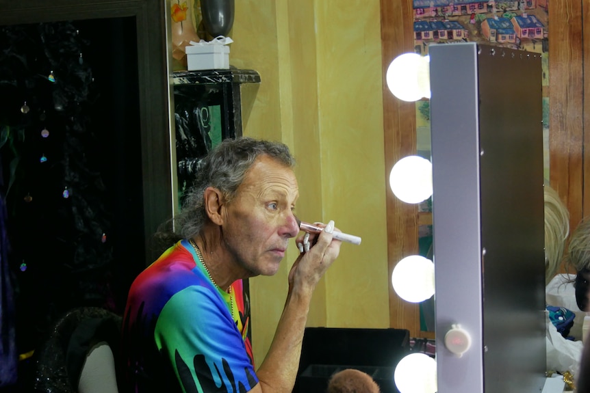 A man sits in front of a mirror surrounded by light bulbs,  brushing a make-up brush across his face. 