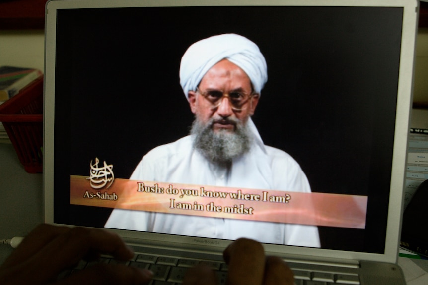 An open laptop displays a full-screen video of a bearded man in white turban speaking, with partial subtitles 