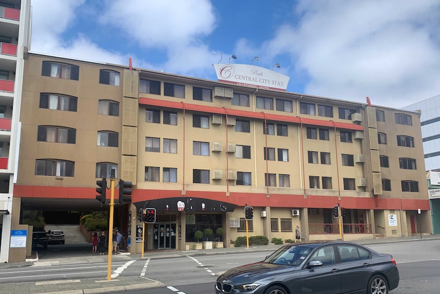 An four-storey inner-city hotel, with creme facade on a street with traffic lights.