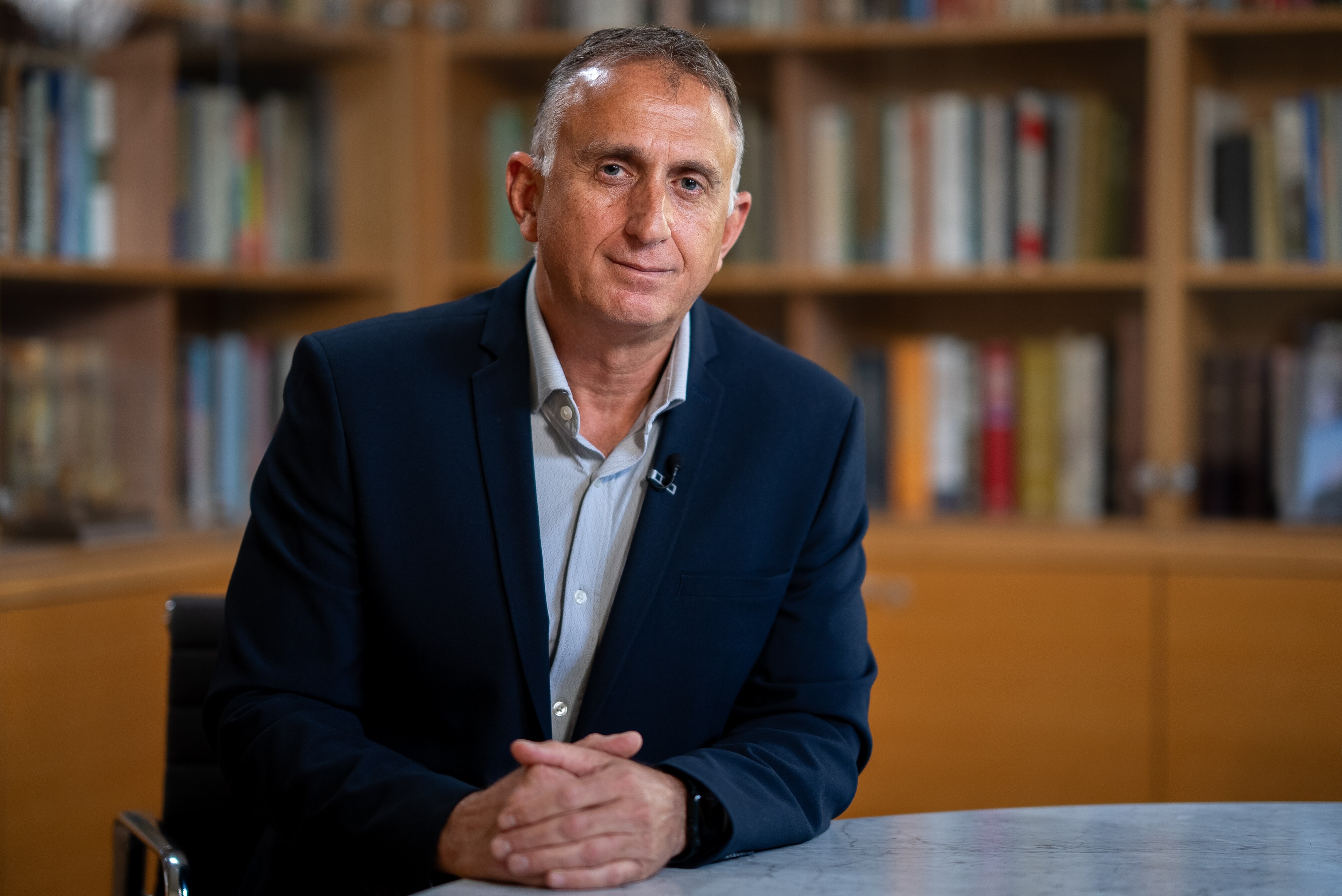 Tamir Hayman wears a blue suit coast as he sits at a desk with his hands together.