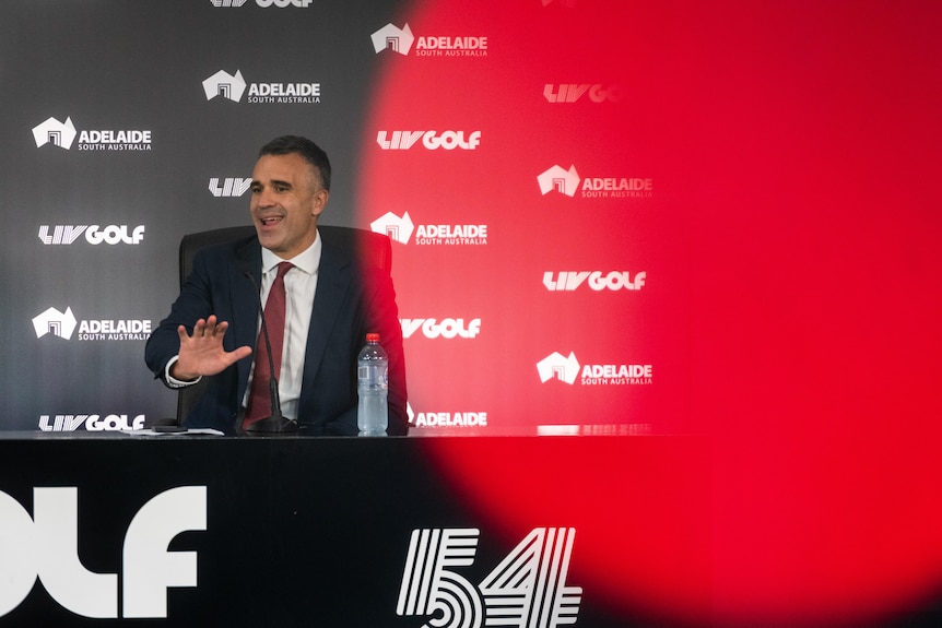 A man in a suit and red tie sitting at a table with LIV Golf signage