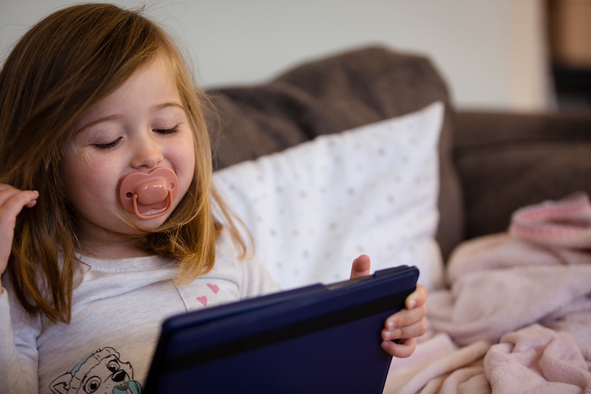Three-year-old Harlow sits on the couch smiling at a show on an iPad.
