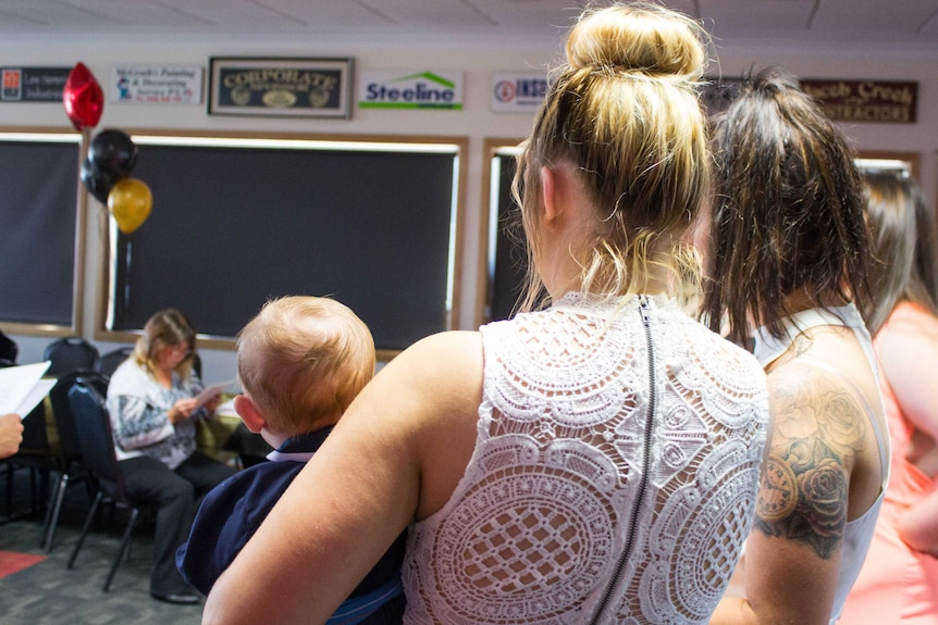 The back of a young mother graduating from high school - wearing a nice dress and holding her baby on her left hip.