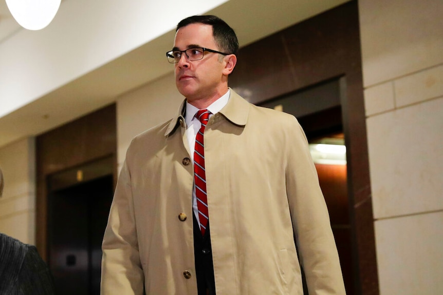 A man in a beige trench coat and red tie walks through a hallway in the American Congress.