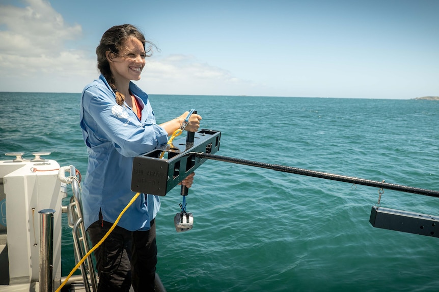 A woman in a blue long-sleeved shirt standing on the side of a boat over the ocean with a large antennae in her hands.