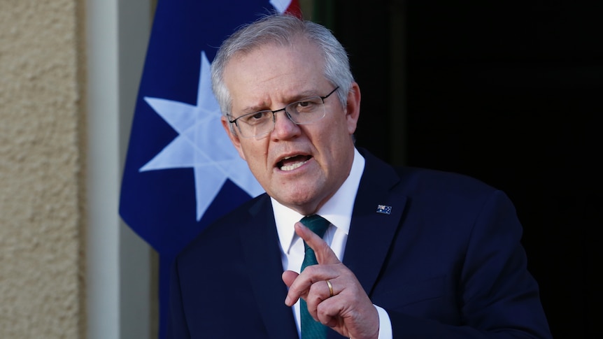 Scott Morrison stands infront of an Australian flag mid-sentence with his finger pointing