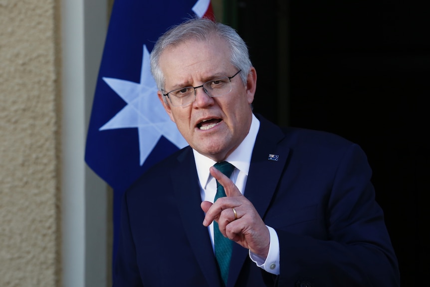 Scott Morrison stands infront of an Australian flag mid-sentence with his finger pointing
