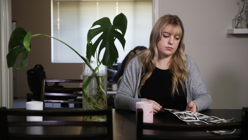A young woman with blonde hair sits at a dining table looking at baby ultrasound pictures, with an indoor plant behind her.