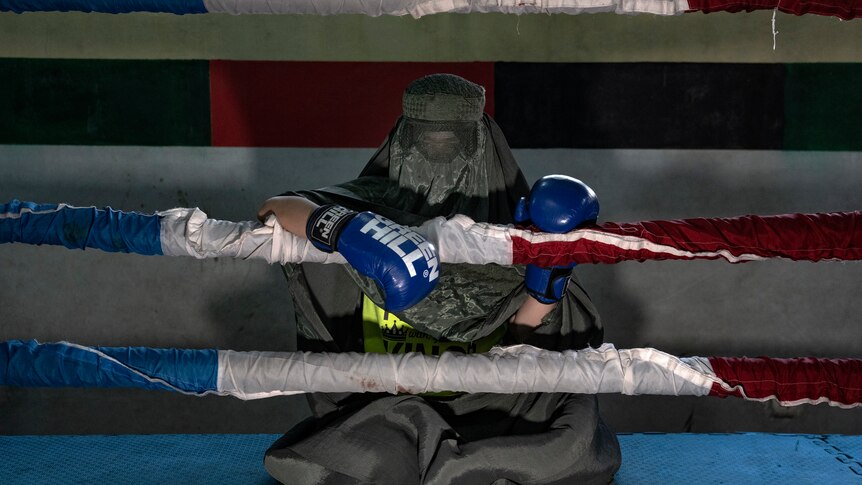 Afghan woman boxer wears a burqa and sits behind the ropes of a boxing ring.