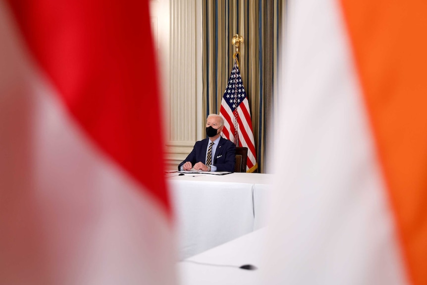 An elderly man in a dark suit and face mask sits at white-tableclothed desk in front of US flag.