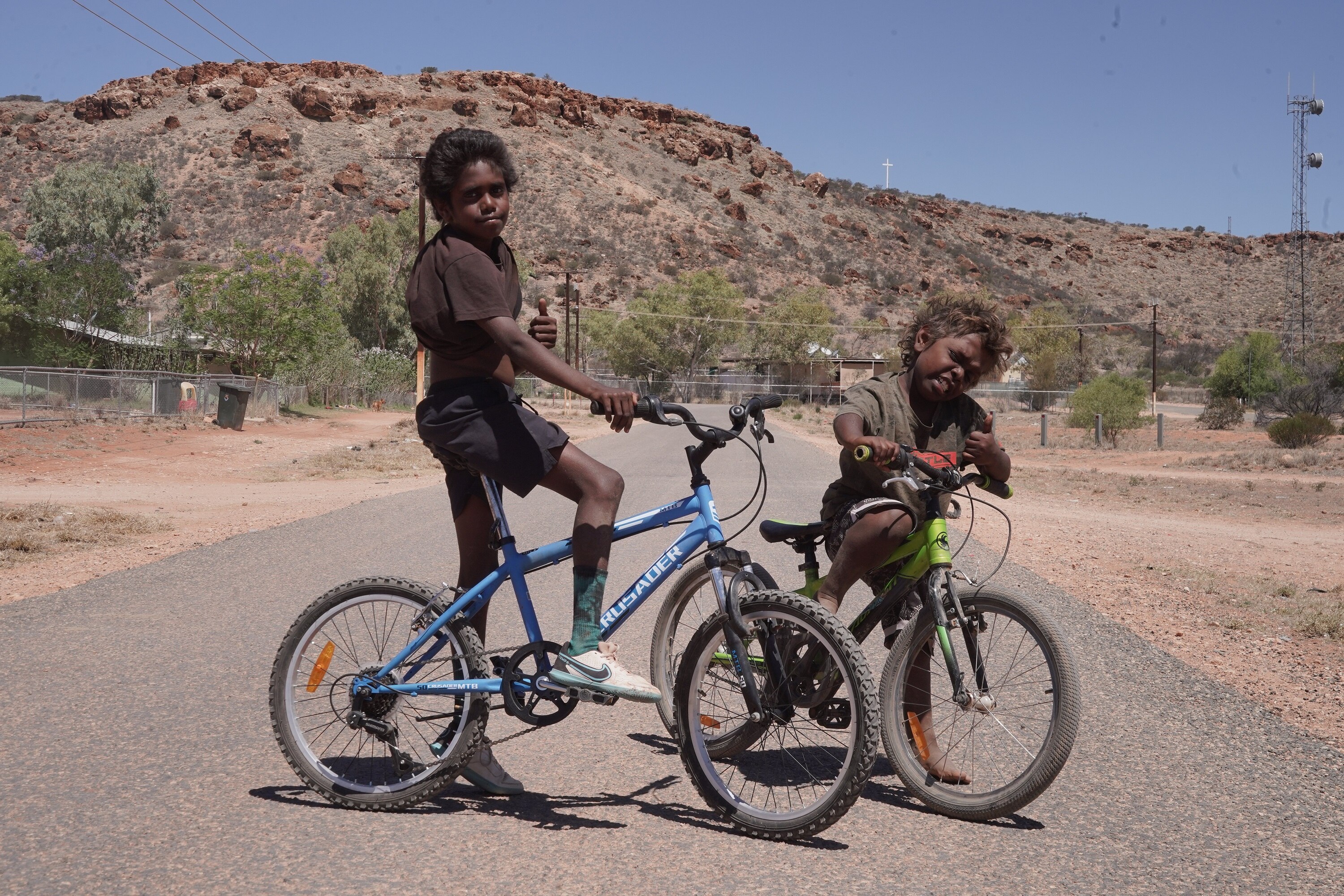 Two Aboriginal children riding bikes on the road near red dirt.