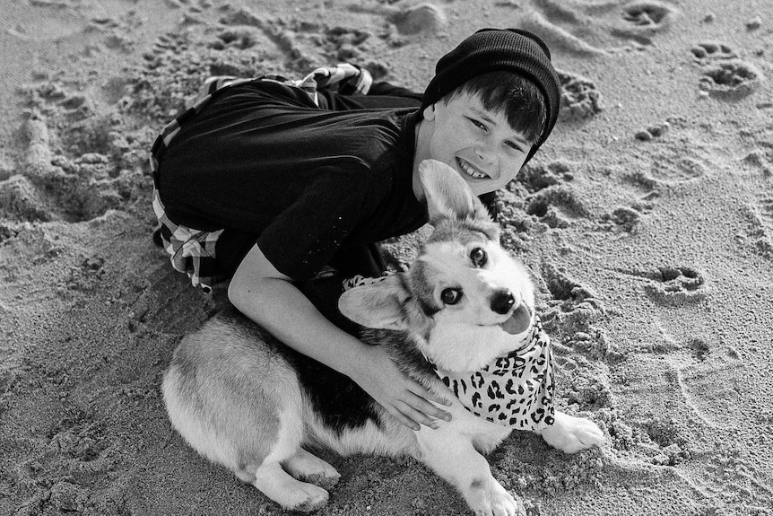 Een jongen en een corgi zitten op het strand, glimlachen en kijken naar de camera.
