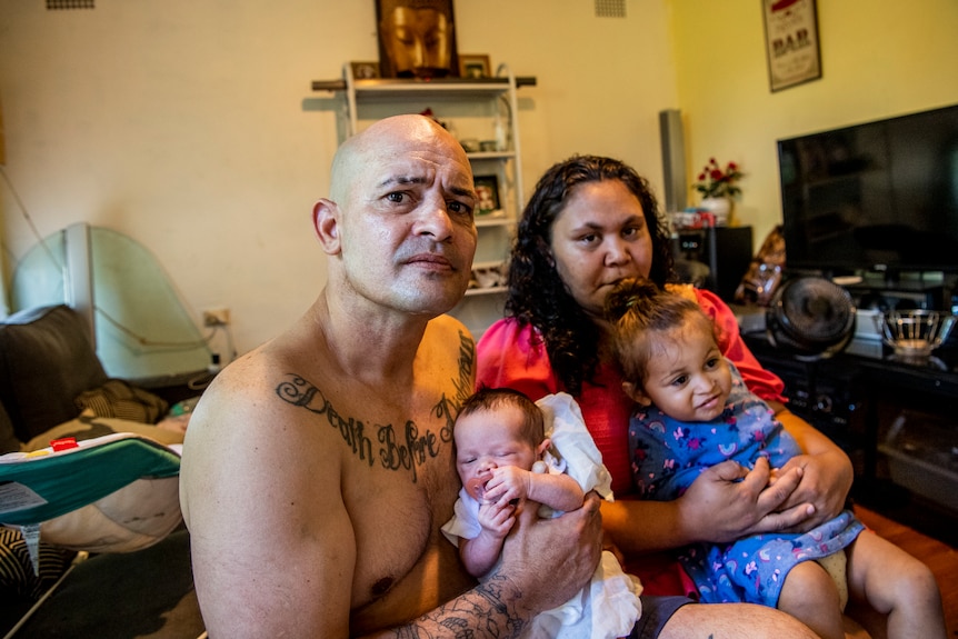 Indigenous man with tattoo across his chest holding a baby, sitting next to his partner and child.