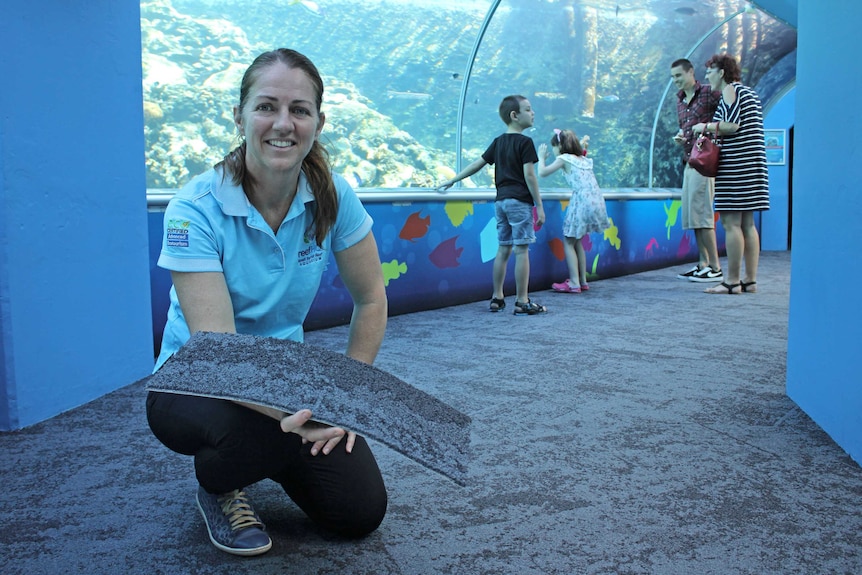 A woman with long brown hair kneels on carpet in front of an aquarium tank.