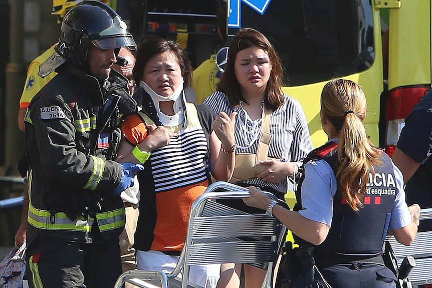 An emergency worker wearing a helmet and a civilian help a woman in a neck brace to walk. Ambulance is in the background.
