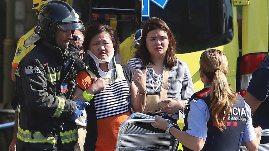 An emergency worker wearing a helmet and a civilian help a woman in a neck brace to walk. Ambulance is in the background.