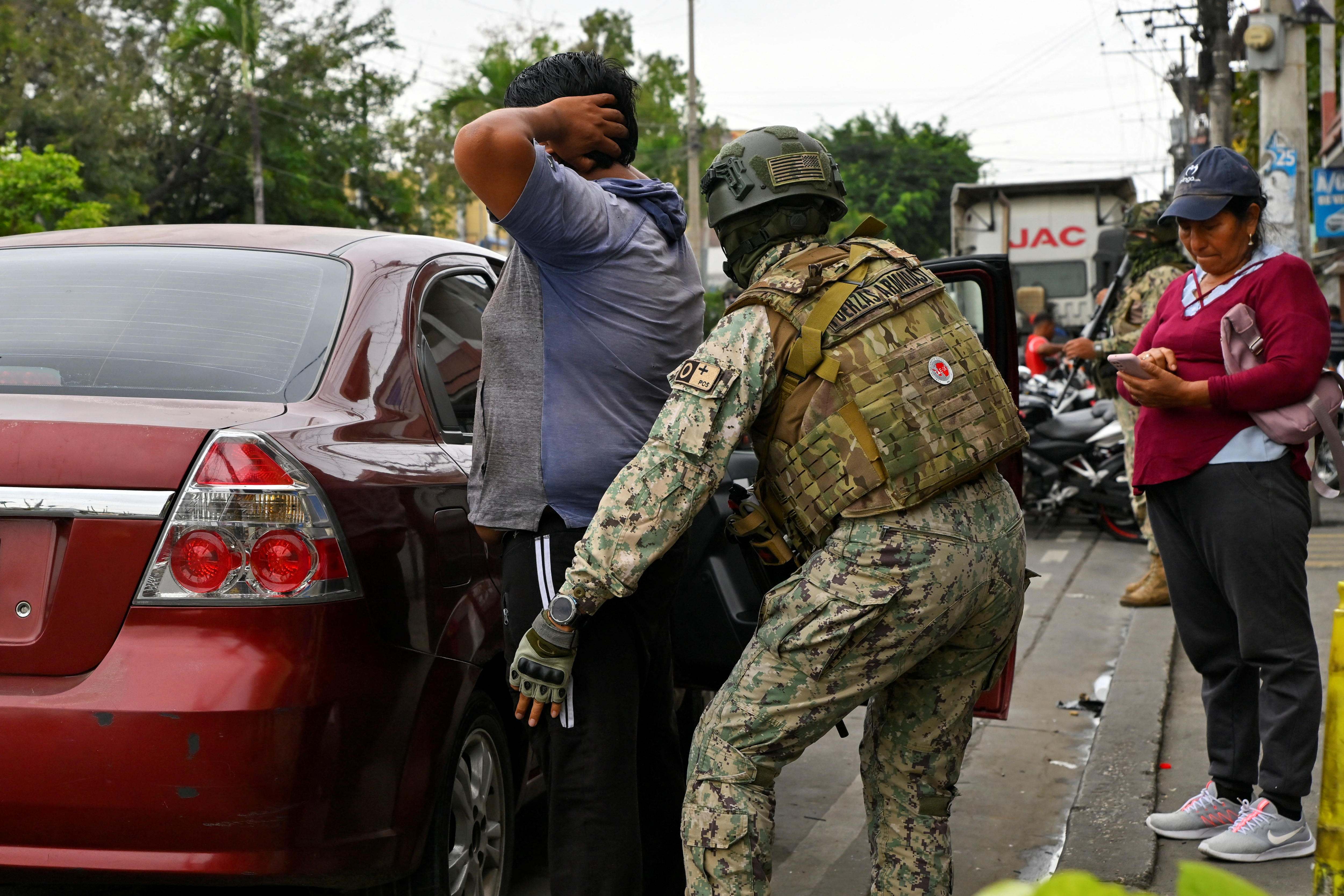 Um oficial militar revista um homem perto de um carro vermelho. 