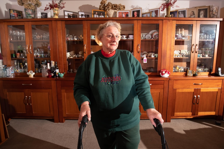 A woman with a walker stands in front of a China cabinet with framed pictures of her family sitting on top.