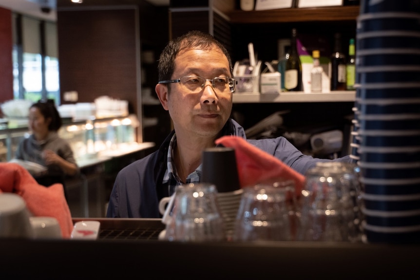 A man stands behind the counter of a cafe, facing the camera but with his eyes looking away