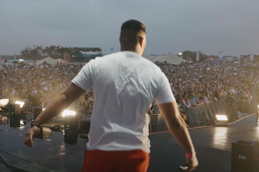 Man's back to camera as crowd of faces look to him on stage. 