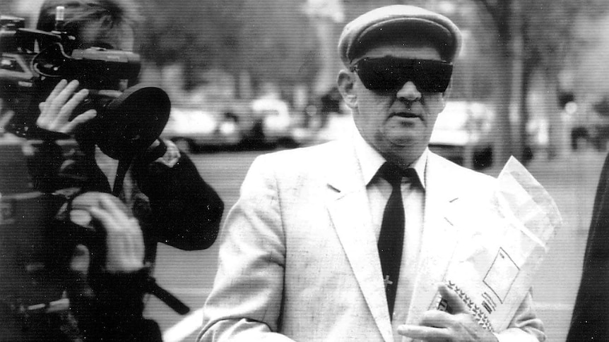 An old man in a driver's cap, large black sunglasses, and a light-coloured suit leave court.