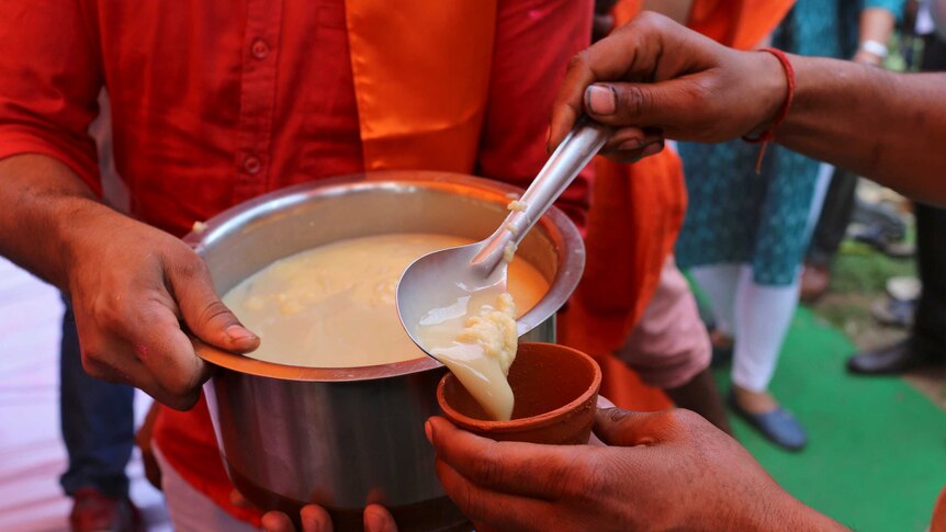 A man holds a pot filled with yellow curdled liquid as a man spoons some of it into a cup.