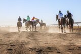 Six horses and jockeys on a dusty race track on a sunny day. Photo shot from the back.