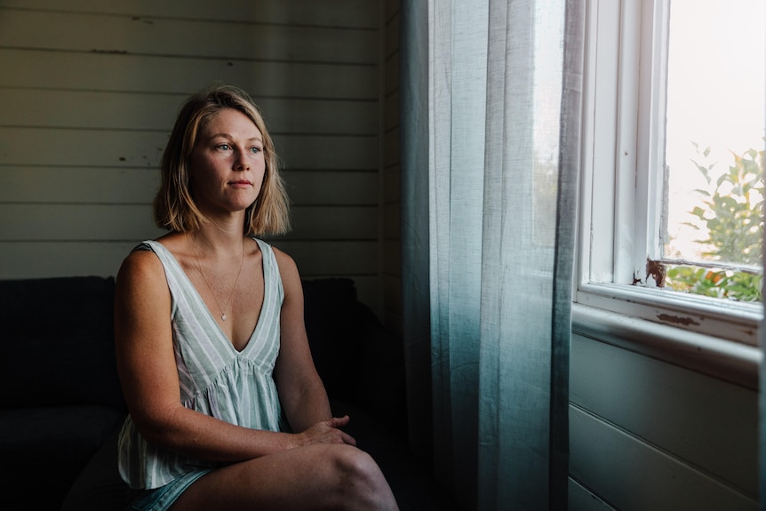 A blonde woman in her thirties in a white top gazes out the window