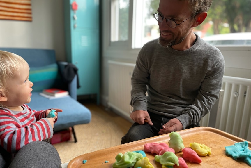 A man and a toddler look at each other smiling in front of a table covered with colourful play-dough.