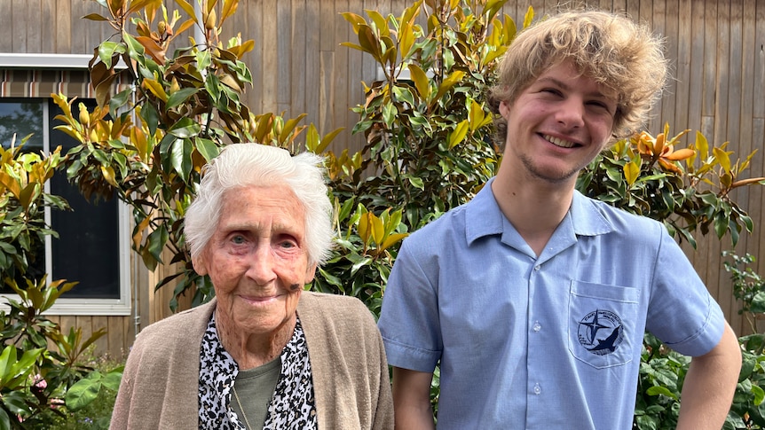 An elderly woman stands with a teenage boy in a garden.