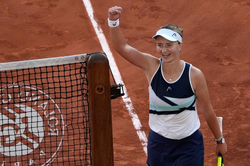 A tennis player smiles and raises her fist in  the air as she stands next to the net after winning a match.