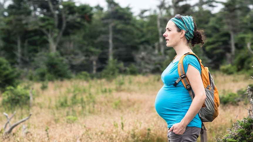Pregnant woman wearing a backpack and hiking gear stands in a wooded area.