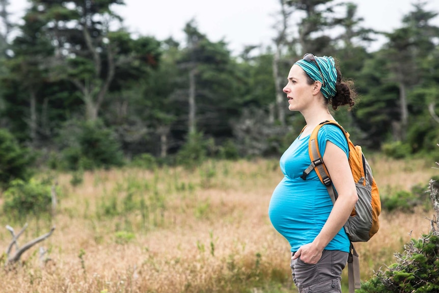 Pregnant woman wearing a backpack and hiking gear stands in a wooded area.