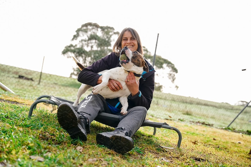 Woman sitting on a dog bed smiling with dog on her lap. Dog looking up at her with a ball in its mouth.